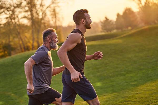 Two men jogging on a grassy hill at sunset, one younger and one older, wearing athletic shorts and shirts.