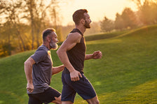 Two men jogging on a grassy hill at sunset, one younger and one older, wearing athletic shorts and shirts.