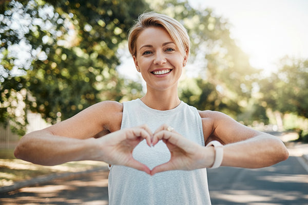 Smiling woman forms a heart shape with her hands while standing outdoors on a sunny tree-lined street.