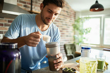 Man adds powder to a shaker bottle beside green smoothies and supplements on a kitchen counter in a bright, sunlit room.
