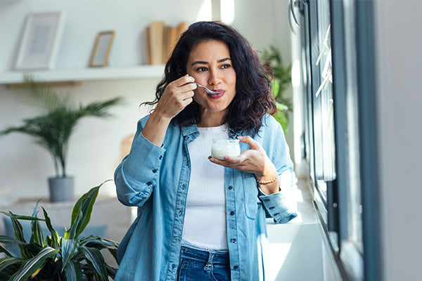 Woman in a blue shirt eats yogurt with a spoon while standing near a sunny window in a bright room filled with houseplants.