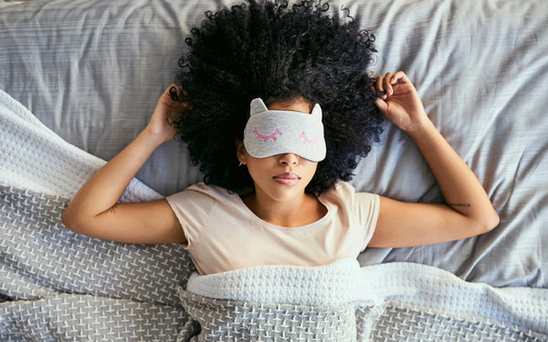 Woman sleeping in bed with her arms up and an eye mask on her face, covering her eyes